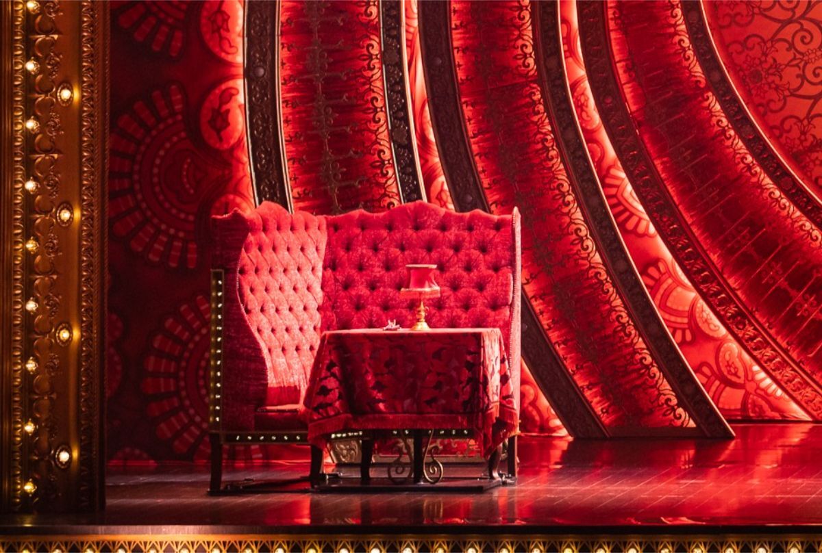 Reveal Productions’ red tufted booth and table with patterned fabric used in Moulin Rouge’s set design, illuminated under stage lighting.