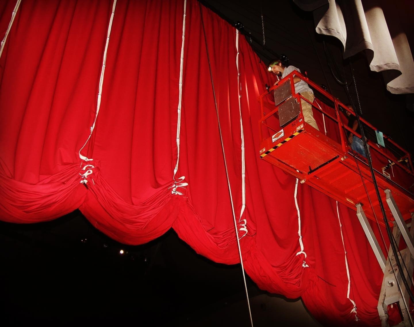 Installer on an elevated platform setting the lift lines of a red venetian curtain above a stage